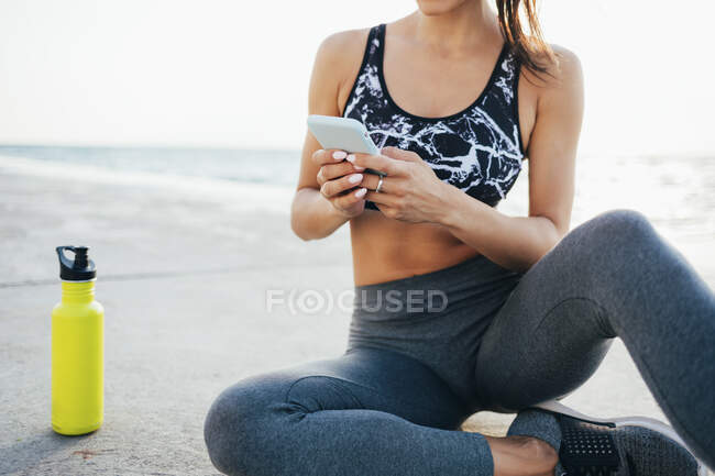 Low section of young woman using mobile phone after exercising on promenade — Stock Photo