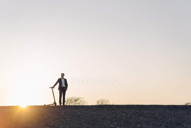 Homem de negócios maduro com uma scooter pontapé em uma ponta de mina desutilizada ao pôr do sol — Fotografia de Stock