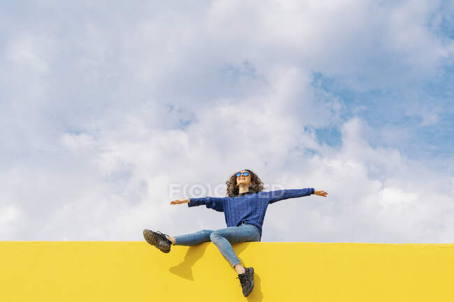 Jovem feliz sentada na parede amarela contra o céu nublado olhando para cima — Fotografia de Stock