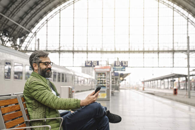 Uomo seduto su una panchina alla stazione ferroviaria utilizzando lo smartphone — Foto stock