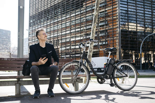 Uomo d'affari dai capelli grigi seduto su una panchina accanto alla bicicletta elettrica in città — Foto stock