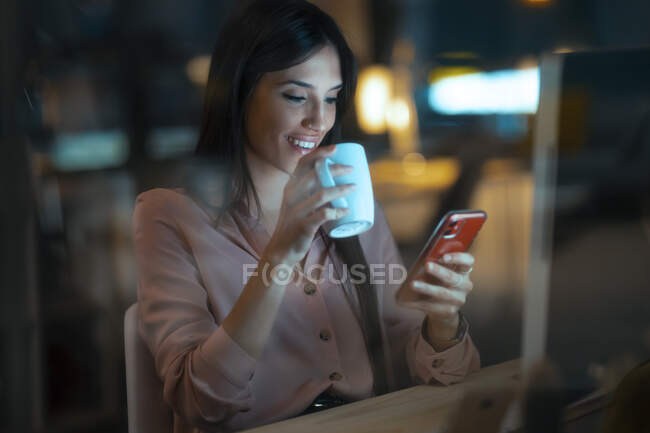 Mujer joven sonriente con taza de café sentado en el escritorio en la oficina mirando el teléfono inteligente - foto de stock