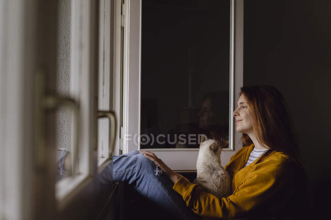 Mujer pelirroja sonriente sentada en la ventana abierta con su gato mirando a la distancia - foto de stock