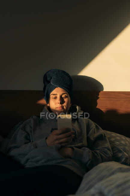 Jeune femme avec la tête enveloppée dans une serviette couchée dans le lit à la maison en utilisant un smartphone — Photo de stock