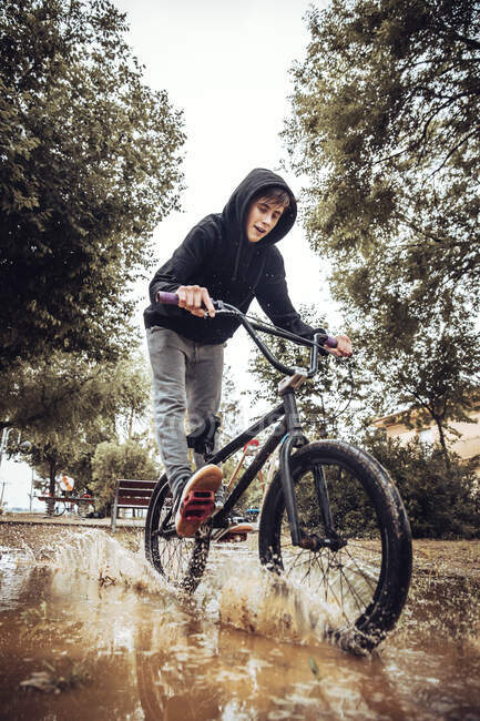 Adolescent garçon éclaboussures eau dans flaque tout en vélo pendant la saison des pluies — Photo de stock