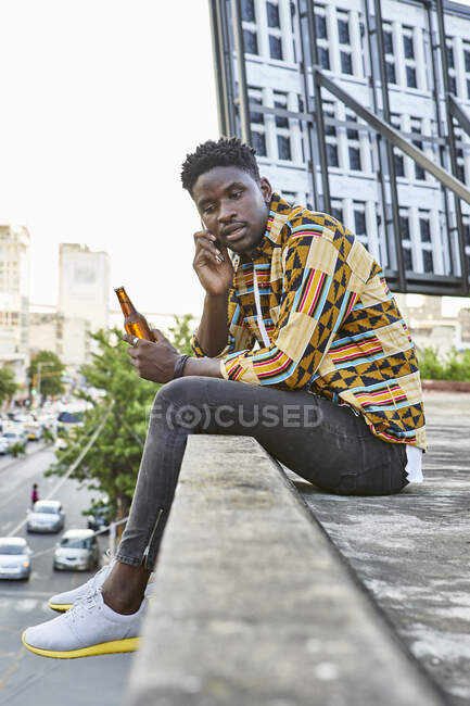 Jeune homme en chemise à motifs assis sur la terrasse du toit dans la ville parlant au téléphone, Maputo, Mozambique — Photo de stock