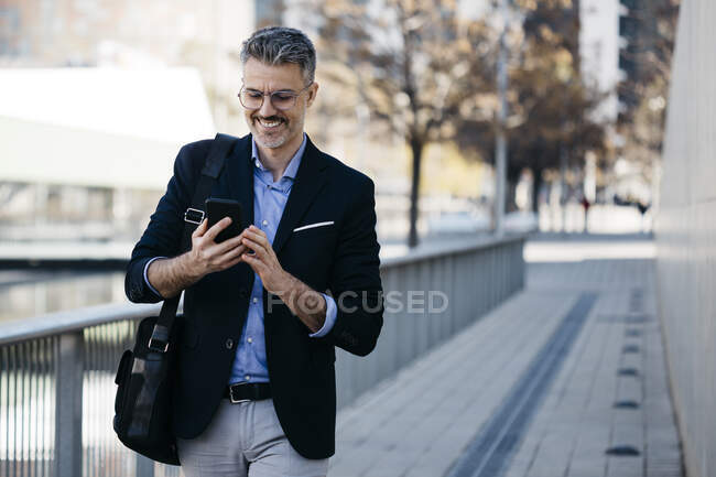 Homme d'affaires souriant aux cheveux gris marchant dans la ville avec un téléphone portable — Photo de stock