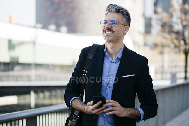Homme d'affaires souriant aux cheveux gris avec téléphone portable dans la ville — Photo de stock