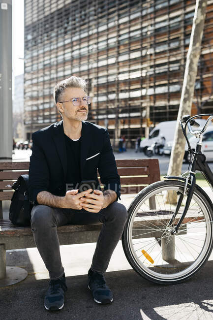 Homme d'affaires aux cheveux gris assis sur un banc à côté du vélo dans la ville — Photo de stock