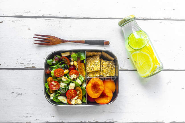 Lunch box with rocket salad with colored tomatoes, mozzarella and nuts, crispbread and apricots and bottle of infused water with lemons and slices of lime — Stock Photo