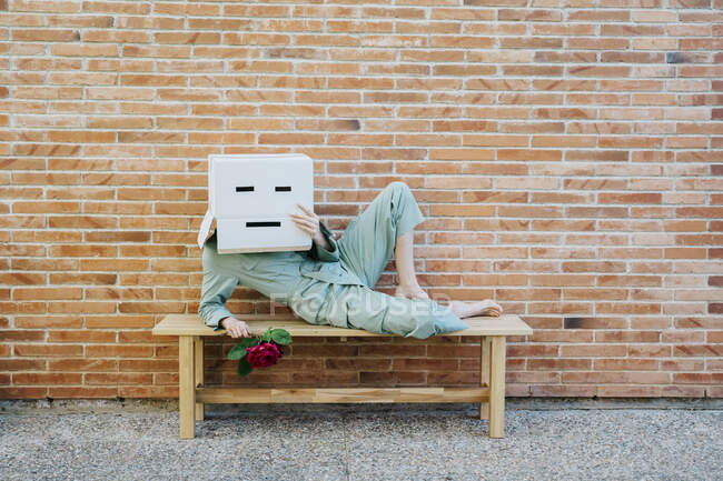 Mujer aburrida con rosa roja, con caja de cartón con cara triste, acostada en el banco frente a la pared de ladrillo - foto de stock