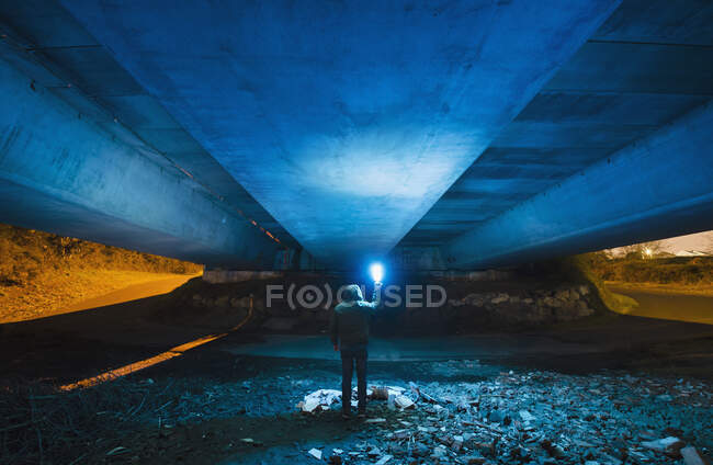 Испания, Галиция, вид сзади на человека в капюшоне, стоящего под бетонным мостом с ярко-голубым светом в руке — стоковое фото