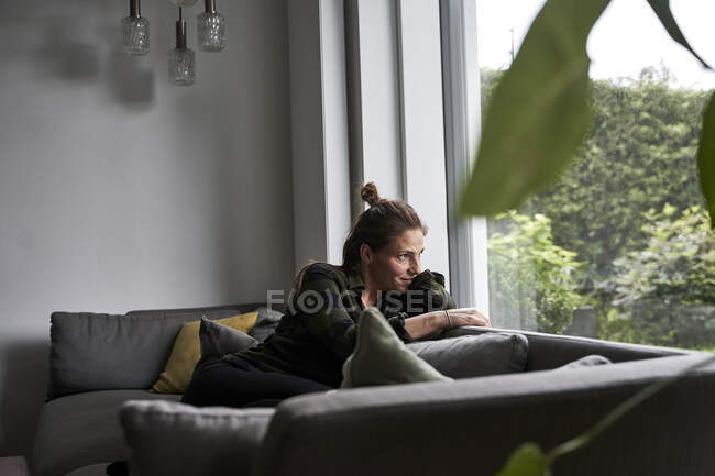 Задумчивая женщина смотрит в окно, расслабляясь дома на диване — стоковое фото
