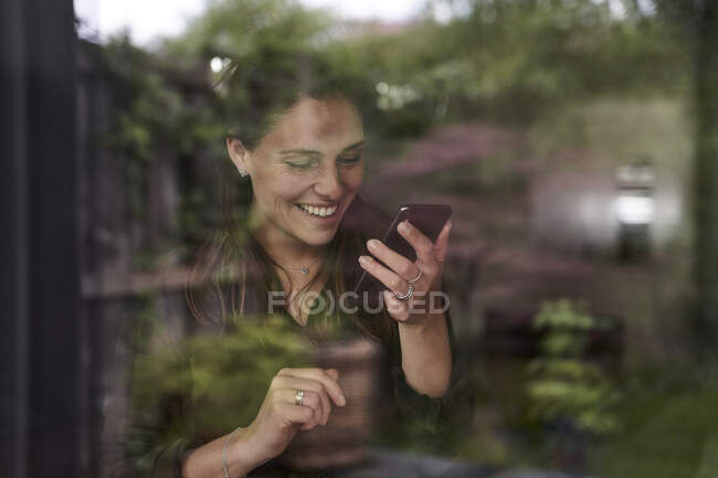 Femme mi-adulte souriante utilisant un téléphone portable à la maison vu par la fenêtre — Photo de stock