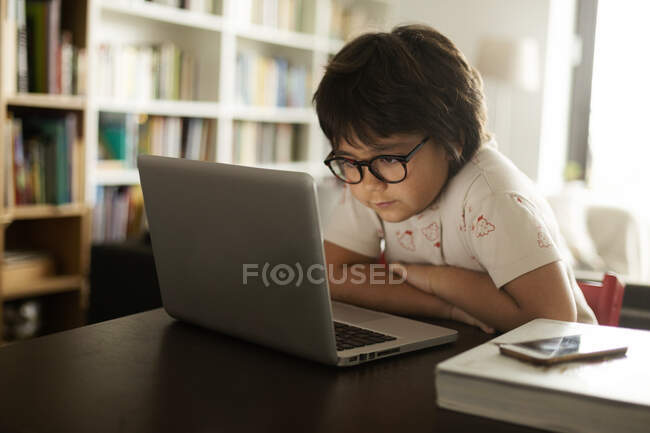 Милий хлопчик з окулярами за допомогою ноутбука, сидячи за столом у вітальні. — стокове фото