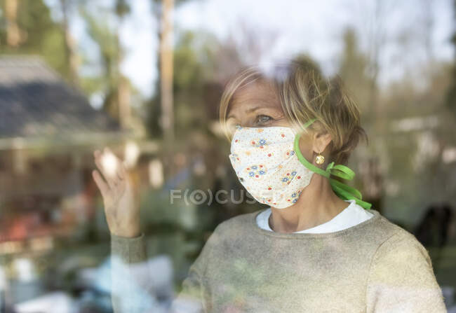 Mulher madura com máscara facial olhando através do vidro da janela durante a pandemia de COVID-19 — Fotografia de Stock