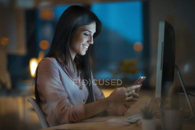 Happy young woman sitting at desk in office looking at smartphone — Stock Photo