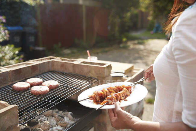 Midsection of woman holding plate with skewers while standing by barbecue grill at back yard on sunny day — Stock Photo