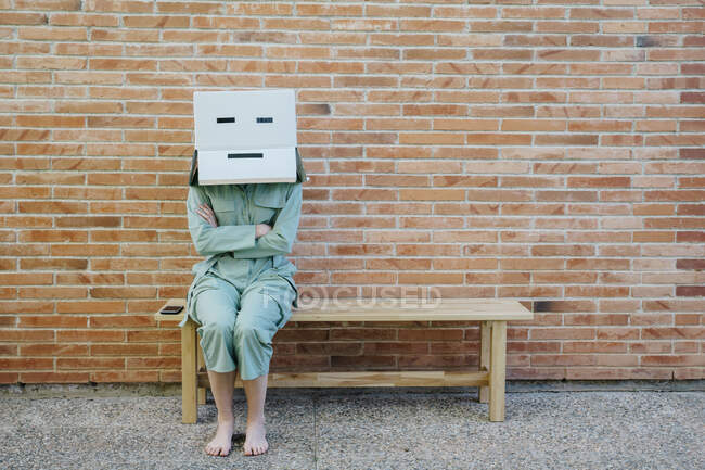 Donna seduta su panchina davanti al muro di mattoni con faccia seria su scatola di cartone, braccia incrociate — Foto stock