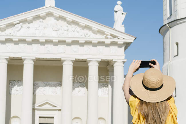 Vista trasera de la mujer fotografiando la catedral con teléfono inteligente en la ciudad durante el día soleado - foto de stock