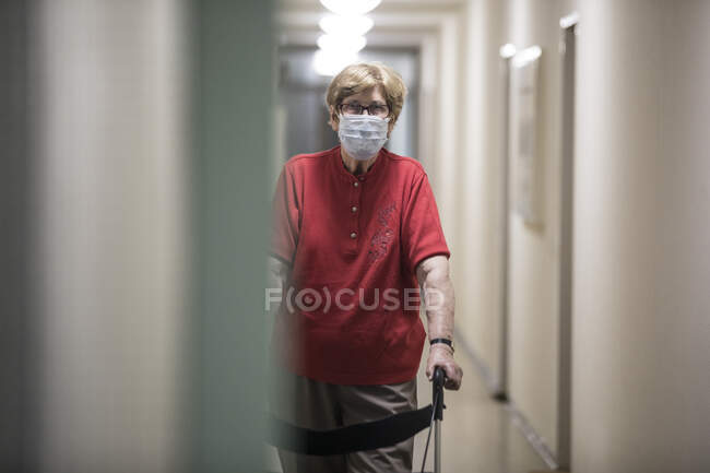Старша жінка в масці і ходить з колісним ходунки в коридорі пенсійного дому — стокове фото