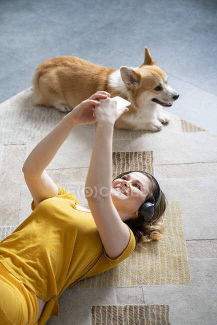 Woman with dog and headphones lying on the floor at home using smartphone — Stock Photo