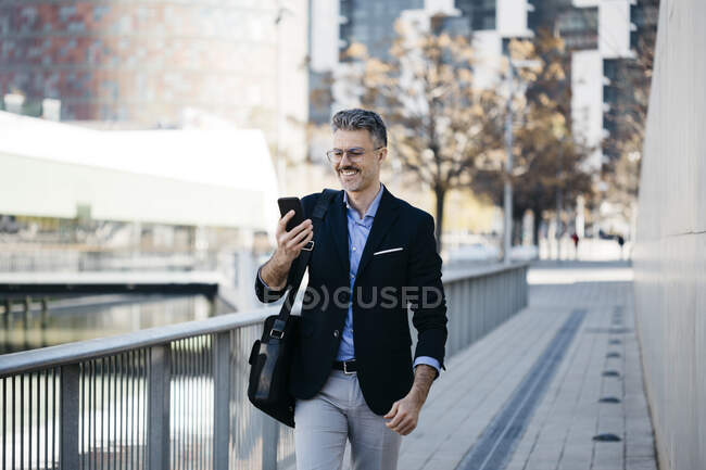 Homme d'affaires souriant aux cheveux gris marchant dans la ville avec un téléphone portable — Photo de stock