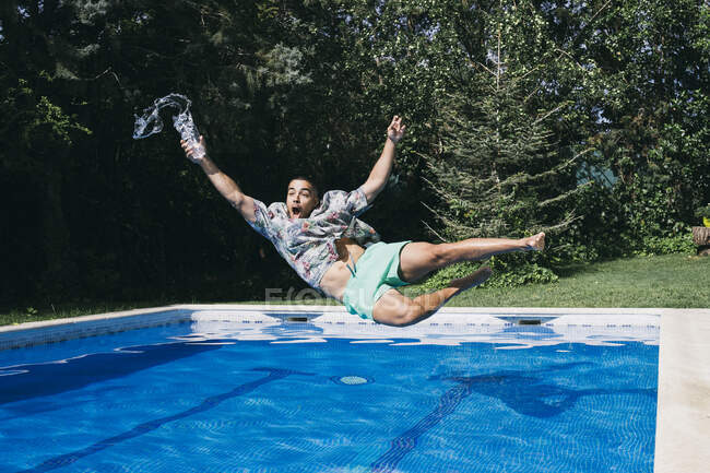 Shocked young man holding drink falling in swimming pool against trees — Stock Photo