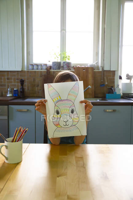 Маленька дівчинка сидить за кухонним столом і тримає малюнок пасхального кролика перед лицем. — стокове фото