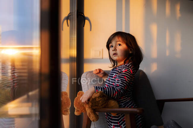 Porträt eines kleinen Mädchens mit Teddybär, das bei Sonnenuntergang aus dem Fenster schaut — Stockfoto