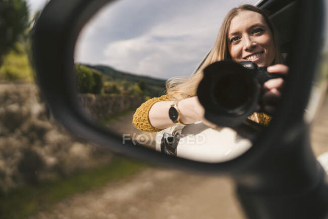 Reflexão no espelho da asa da jovem sorridente com câmera inclinada para fora da janela do carro — Fotografia de Stock
