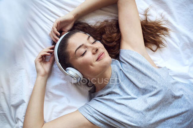 Портрет щасливої молодої жінки з закритими очима лежить на ліжку слухаючи музику з навушниками — стокове фото
