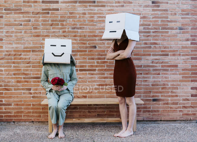 Persona con la cara feliz en la caja de cartón sentado en el banco, dando rosas rojas a la mujer triste - foto de stock
