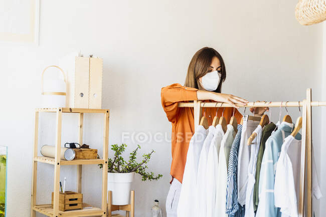 Designer de moda feminina trabalhando em casa com roupas stand usando máscara protetora — Fotografia de Stock