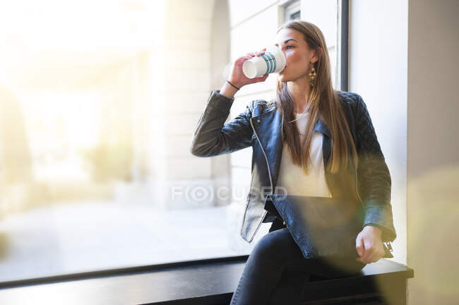 Giovane donna che beve caffè tenendo il computer portatile sul davanzale della finestra in caffetteria — Foto stock