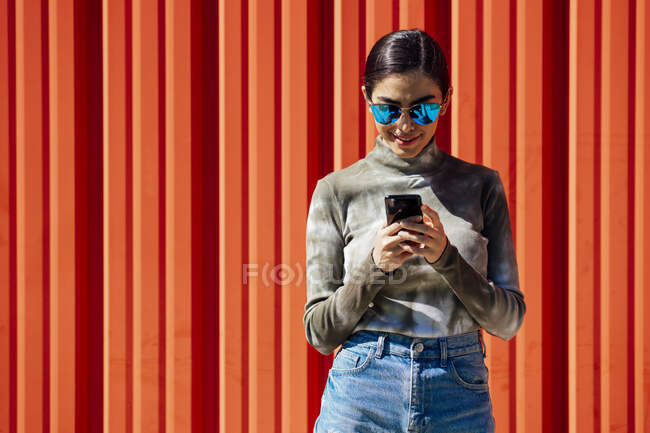 Sorrindo jovem mulher usando telefone inteligente, enquanto em pé contra a parede ondulada vermelha no dia ensolarado — Fotografia de Stock