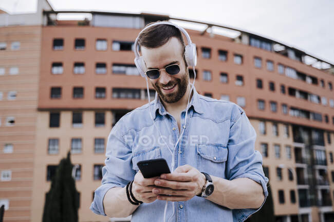 Retrato de un joven sonriente con auriculares mirando el teléfono celular - foto de stock