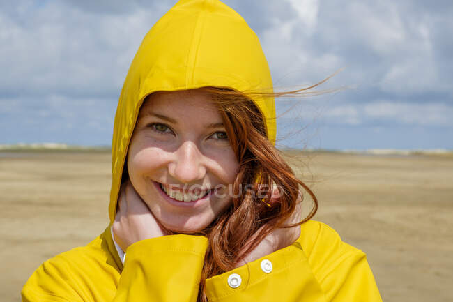Close-up portrait of redhead teenage girl wearing yellow raincoat while standing at beach against sky on sunny day — Stock Photo