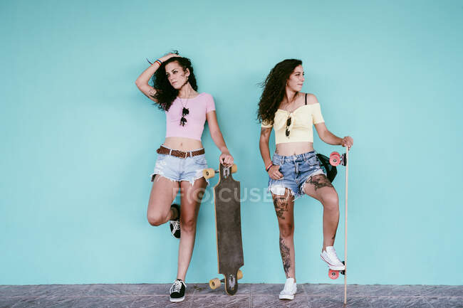 Casal feminino olhando para longe enquanto estava de pé com skates contra a parede azul na cidade — Fotografia de Stock