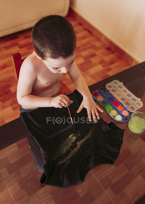 Vue grand angle de garçon torse nu dessinant sur t-shirt avec des peintures à l'aquarelle à la maison — Photo de stock