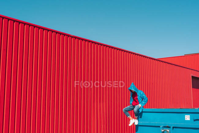 Jeune homme vêtu d'un imperméable assis sur le bord du conteneur bleu devant le mur rouge, regardant vers le bas — Photo de stock