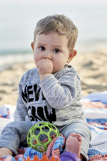 Милий хлопчина смокче великий палець, сидячи на ковдрі на пляжі. — стокове фото