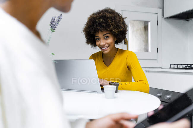 Retrato de sorrindo jovem mulher sentada na cozinha com seu amigo usando laptop — Fotografia de Stock