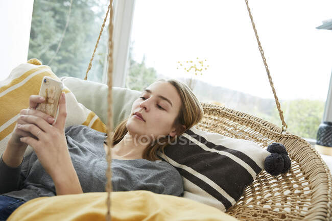 Retrato de una mujer joven acostada en el columpio en casa usando un teléfono inteligente - foto de stock