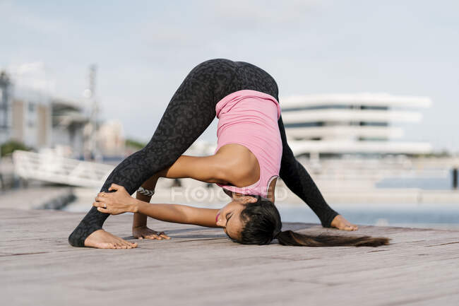 Female athlete practicing wide-legged forward bend on pier against sky — Stock Photo