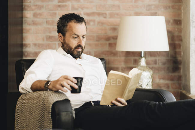 Mature man sitting in armchair, reading book, drinking coffee — Stock Photo