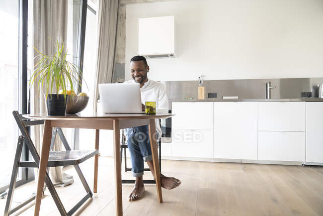 Ritratto di uomo sorridente seduto a tavola in appartamento moderno con laptop e auricolari — Foto stock