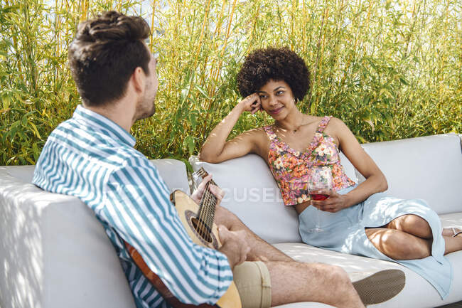 Frau hält Rotweinglas in der Hand, während sie Mann beim Gitarrespielen auf Sofa im Penthouse-Patio beobachtet — Stockfoto