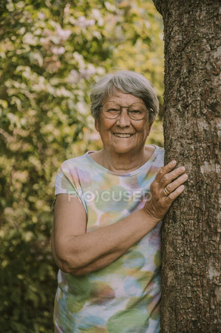 Feliz anciana jubilada de pie junto al árbol contra las plantas en el jardín - foto de stock