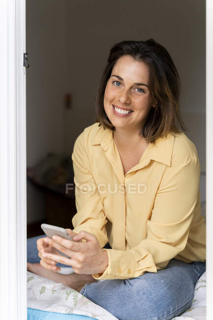 Mulher sorridente com telefone celular sentado no quarto — Fotografia de Stock